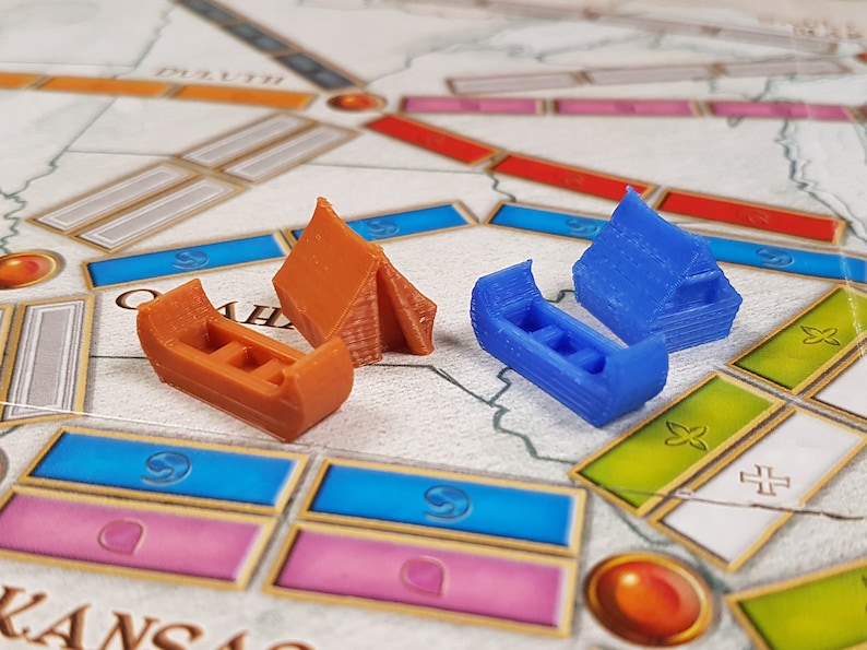 May include: Two miniature wooden houses, one blue and one brown, are placed on a board game with a map of the United States. The houses are made of wood and have a simple design.
