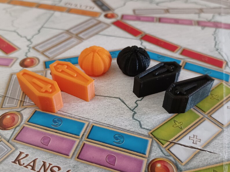 May include: A set of game pieces for a board game. The pieces include two orange coffins, two black coffins, a black sphere, and an orange sphere.