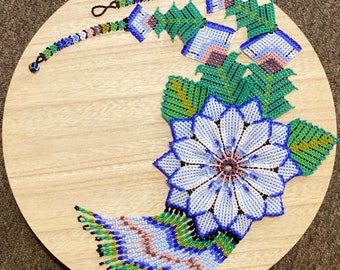 May include: A handcrafted beaded floral design in blue, white, green, and pink hues, arranged on a round wooden surface. The design includes a large flower, leaves, and a beaded bracelet. The intricate beadwork showcases a detailed and colorful pattern.