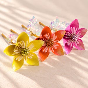 May include: Three decorative hair pins featuring layered flower designs in yellow, orange, and pink. Each flower has a central cluster of sparkling rhinestones and iridescent, translucent petal accents attached to a thin gold-toned metal pin.