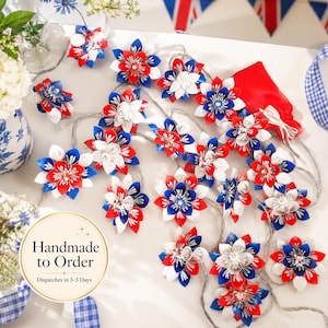 May include: A string of LED fairy lights featuring multi-layered fabric flower ornaments in red, white, and blue. Each flower has pointed petals and a central cluster of small beads. The lights are arranged on a thin, flexible silver wire.