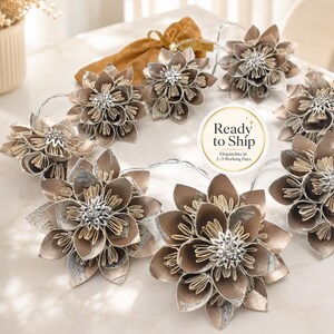 May include: A string of decorative lights featuring multiple layered paper flowers in shades of metallic bronze, gold, and silver. Each flower has a central cluster of clear, sparkling crystals. A gold fabric drawstring pouch is included.