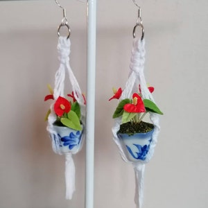 May include: A pair of white macrame earrings with a small blue and white ceramic planter hanging from each. The planters contain a small green plant with red flowers.