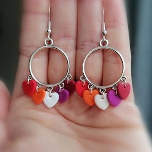 Könnte beinhalten: Ein Paar silberne Creolen mit einem Anhänger aus sechs Herzen in den Farben der lesbischen Pride-Flagge: Rot, Orange, Gelb, Weiß, Rosa und Lila.
