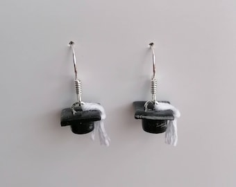 May include: A pair of silver dangle earrings with black graduation cap charms and white tassel accents.