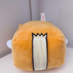 May include: A square-shaped plush toy with soft golden-yellow fabric. The center features a vertical cream-colored stripe bordered by dark outlines, topped with a black crown-like emblem. A small white fabric tag with red text is visible at the top.