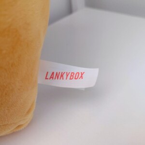 May include: A soft, golden-yellow plush toy featuring a white fabric tag attached to the side. The tag is printed with the text LANKYBOX in bold, red, sans-serif capital letters. The plush material has a smooth, uniform texture.