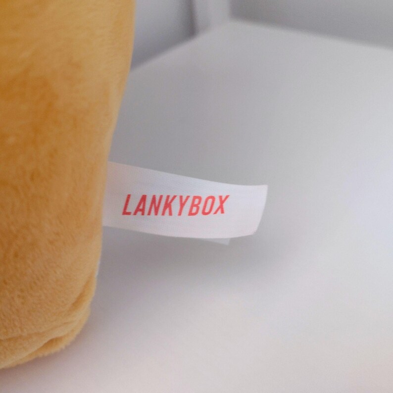 May include: A soft, golden-yellow plush toy featuring a white fabric tag attached to the side. The tag is printed with the text LANKYBOX in bold, red, sans-serif capital letters. The plush material has a smooth, uniform texture.