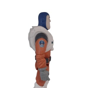 May include: A Buzz Lightyear action figure in a side profile view. The figure is wearing a blue helmet, a white space suit with a black and orange emblem, and orange pants. The figure has gray gloves and white boots.