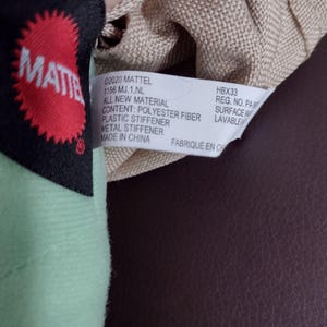 Op de afbeelding: Een beige stoffen tas met een rood en zwart label met de tekst "MATTEL" en een wit label met de tekst "2020 MATTEL 1186 MJ. 1.NL ALL NEW MATERIAL CONTENT: POLYESTER FIBER PLASTIC STIFFENER METAL STIFFENER MADE IN CHINA FABRIQUE EN CH HBX33 REG. NO. PA-887 SURFACE WASHABLE"