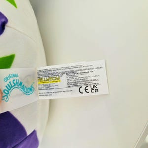 May include: A white plush toy with purple and green accents. A tag is attached with the words "Original Squishmallows" and "Kellytoy". The tag also includes the company's name and address.