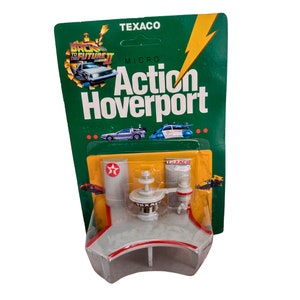 May include: A white plastic model of a hoverport from the film Back to the Future II. The hoverport is a futuristic petrol station with a Texaco logo and a red and white striped awning. The model is packaged in a green and yellow cardboard box with the words "Micro Action Hoverport" printed on it.