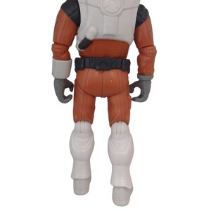 May include: A toy action figure of a space explorer, viewed from the back. The figure is wearing an orange jumpsuit with white accents on the chest, boots, and cuffs. The figure has gray gloves and a black belt. The figure is standing.