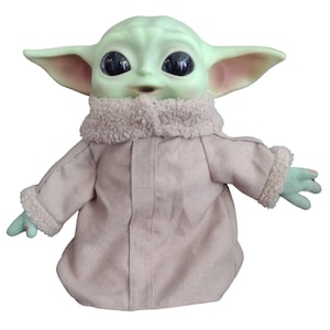 Op de afbeelding: Een pluche speelgoed van het personage Baby Yoda uit de Star Wars-serie The Mandalorian. Het speelgoed draagt een bruine jas met een witte bontkraag en manchetten. Het speelgoed heeft grote ogen en een groene huidskleur.