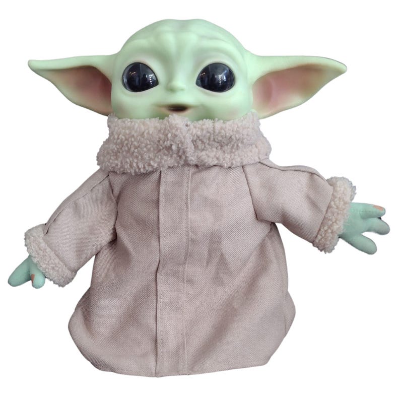 Op de afbeelding: Een pluche speelgoed van het personage Baby Yoda uit de Star Wars-serie The Mandalorian. Het speelgoed draagt een bruine jas met een witte bontkraag en manchetten. Het speelgoed heeft grote ogen en een groene huidskleur.