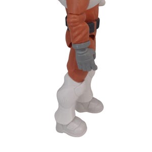 May include: A toy action figure of an astronaut in a white and orange spacesuit. The figure has gray gloves, a black belt, and white boots. The figure is standing on a white surface.