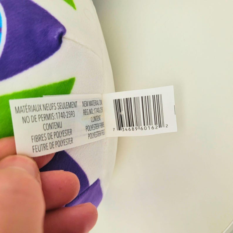 May include: A white plush toy with a colorful design of purple, green, and blue shapes. A tag is attached with text in French and English, including "NEW MATERIAL ON" and "POLYESTER FIBRE".