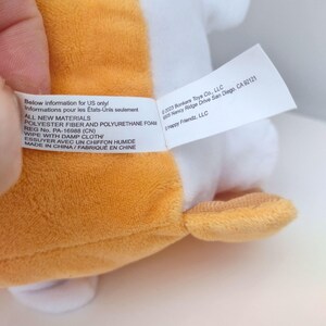 May include: A plush toy with orange and white fabric featuring a white care label with black text. The label reads: Below information for US only, ALL NEW MATERIALS, POLYESTER FIBER AND POLYURETHANE FOAM, REG No. PA-16988 (CN), WIPE WITH DAMP CLOTH, MADE IN CHINA, 2023 Bonkers Toys Co., LLC, 6905 Nancy Ridge Drive San Diego, CA 92121, Happy Friendz, LLC.