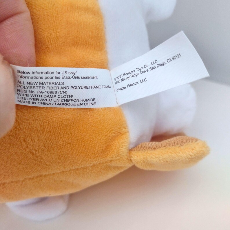 May include: A plush toy with orange and white fabric featuring a white care label with black text. The label reads: Below information for US only, ALL NEW MATERIALS, POLYESTER FIBER AND POLYURETHANE FOAM, REG No. PA-16988 (CN), WIPE WITH DAMP CLOTH, MADE IN CHINA, 2023 Bonkers Toys Co., LLC, 6905 Nancy Ridge Drive San Diego, CA 92121, Happy Friendz, LLC.