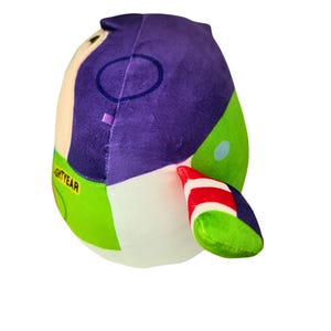 May include: A plush toy shaped like Buzz Lightyear from Toy Story. The toy features a purple head, green and white accents, and the word "LIGHTYEAR" printed on the side. The toy is designed with a rounded shape.