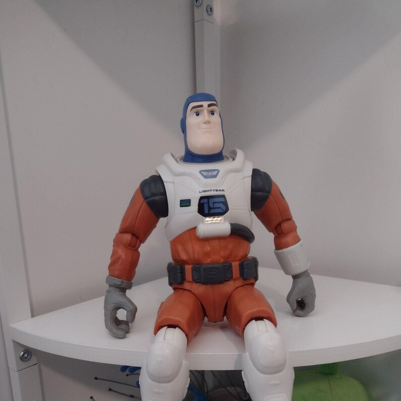 May include: A Buzz Lightyear action figure, featuring a white and orange spacesuit with the number 15 on the chest. The figure has a blue helmet and gloves, and is posed sitting down. The toy is a collectible item.