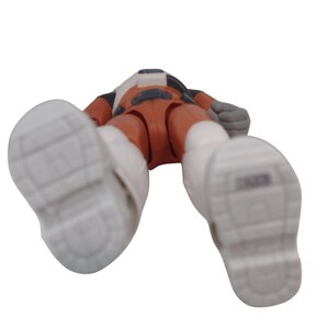 May include: A toy action figure in an orange and white spacesuit, lying on its back. The figure has white boots and gray gloves. The figure's head is turned away from the camera.