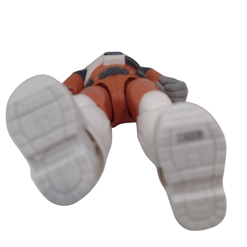 May include: A toy action figure in an orange and white spacesuit, lying on its back. The figure has white boots and gray gloves. The figure's head is turned away from the camera.