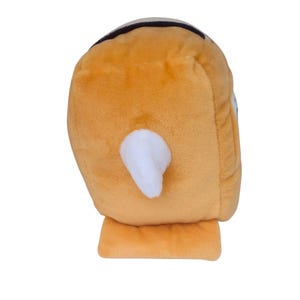 May include: A plush toy shaped like a rounded, golden-yellow character with a soft, fuzzy texture. It features a prominent, triangular white protrusion on its side and a dark, curved detail at the top, presenting a simple, three-dimensional form.