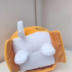 May include: A plush toy featuring a bright orange rounded body with a white rectangular front panel. Two small white rounded feet are attached to the bottom of the white panel. A white fabric tag extends from the top, and the word Boxy is printed.