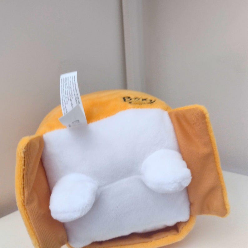 May include: A plush toy featuring a bright orange rounded body with a white rectangular front panel. Two small white rounded feet are attached to the bottom of the white panel. A white fabric tag extends from the top, and the word Boxy is printed.