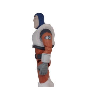 May include: A side view of a Buzz Lightyear action figure. The figure is wearing a blue helmet, white chest armor, and an orange and white spacesuit. The figure has gray gloves and a gray belt. The letters "SC" are printed on the right shoulder.