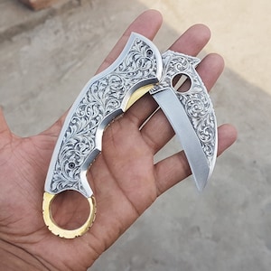Könnte beinhalten: Ein silbernes Karambit-Messer mit aufwendigen floralen Gravuren. Die gebogene Klinge ist teilweise geöffnet und zeigt eine scharfe Schneide. Ein goldfarbener Fingerring befindet sich am Griff. Das Messer wird in einer Hand gehalten.