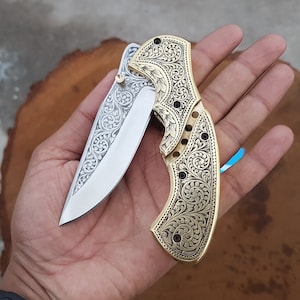 May include: A gold pocket knife with an intricate floral design on the handle and a silver blade. The knife is open and the blade is visible.