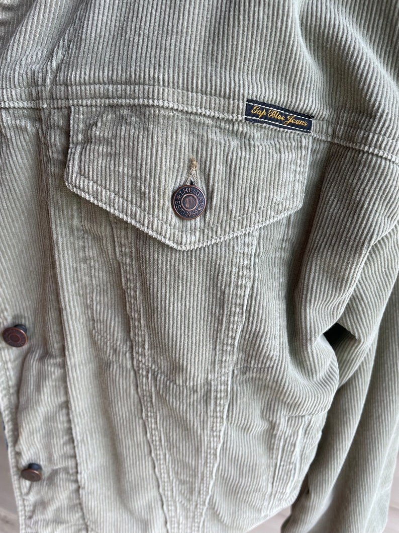 Op de afbeelding: Een lichtgroen corduroy jack met knoopsluiting en een patch op de zak met de tekst "Gap Blue Jeans".