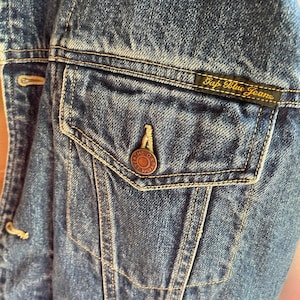 Op de afbeelding: Close-up van een blauw denim jack met een zak en een Gap Blue Jeans label. De zak heeft een kleine metalen knoop met de tekst "The Gap Est. 1969".