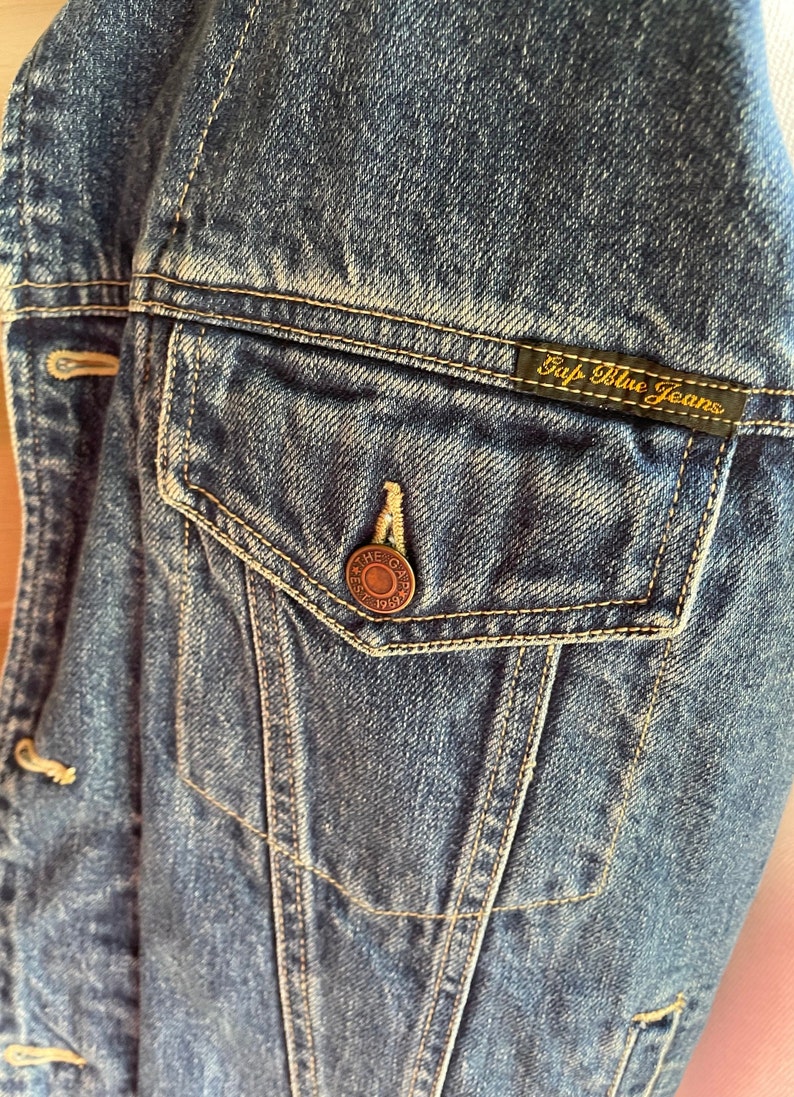 Op de afbeelding: Close-up van een blauw denim jack met een zak en een Gap Blue Jeans label. De zak heeft een kleine metalen knoop met de tekst "The Gap Est. 1969".