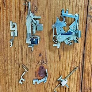 May include: A set of six silver sewing machine feet, including a zig-zag foot, a blind stitch foot, a zipper foot, a buttonhole foot, a straight stitch foot, and a foot with a small white knob.