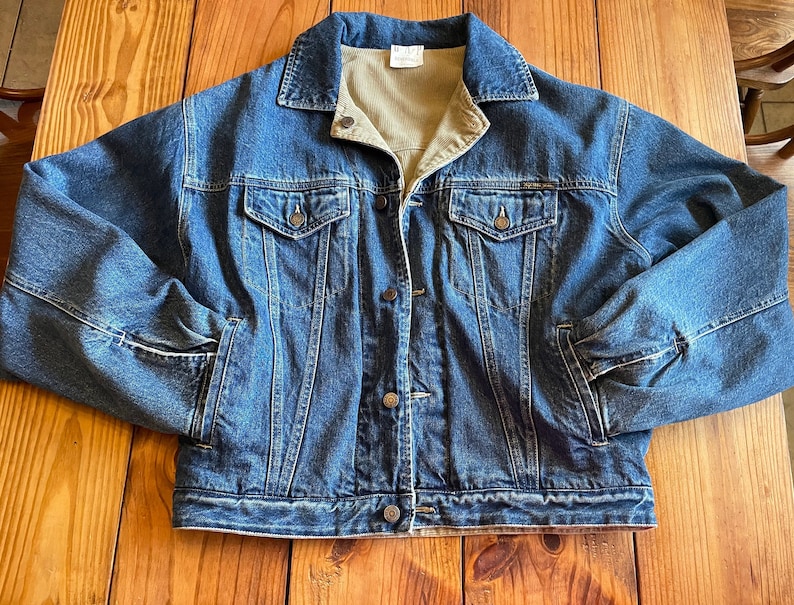 Op de afbeelding: Een blauwe denimjas met een corduroy kraag en knoopsluiting. De jas heeft twee voorzakken en twee borstzakken.