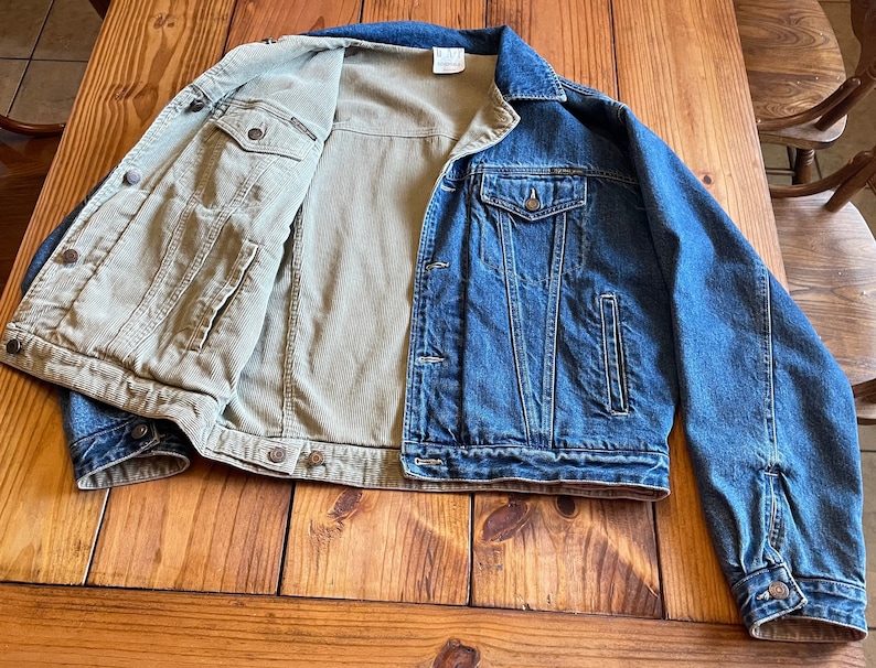 Op de afbeelding: Een spijkerjack met een corduroy voering. Het jack is blauw en de voering is beige. Het jack ligt plat op een houten oppervlak.