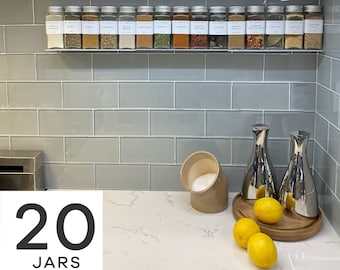 May include: A kitchen countertop with a white countertop and gray subway tile backsplash. A clear acrylic spice rack with 20 glass spice jars is mounted on the backsplash. The jars are labeled with white text. The text "20 JARS" is printed in black on a white background in the lower left corner of the image.