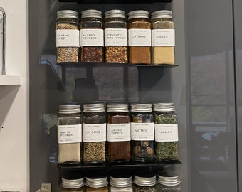 May include: Twenty glass spice jars with black lids and white labels are organized on four black metal shelves. The labels list the spices, including bay leaf, Sichuan pepper, cinnamon stick, paprika, cumin, sesame seed, Aleppo peppers, crushed red pepper, cayenne, ginger, white pepper, oregano, ancho pepper, nutmeg, parsley, garam masala, cinnamon, garlic powder, basil, and onion powder.