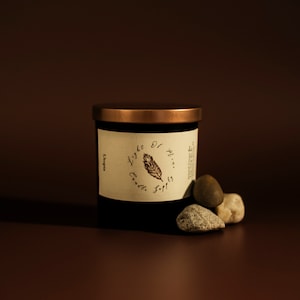May include: A black candle jar with a copper-colored lid and a label with the text "Light of Candle Soap". Several small, smooth stones are arranged near the base of the jar. The background is a dark brown.