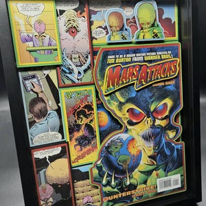 May include: Framed comic book art featuring panels from "Mars Attacks." The artwork includes colorful illustrations of aliens, explosions, and comic book text. The frame is black, and the art is layered for a 3D effect.