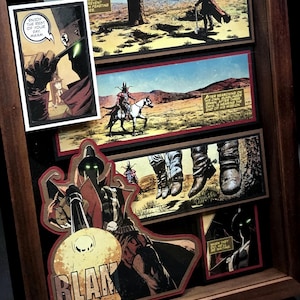 May include: A wooden shadow box frame containing layered comic book panels featuring a gunslinger character. The artwork displays desert landscapes, a figure on horseback, and close-ups of boots and a gun firing with the text BLAM and ENJOY THE REST OF YOUR DAY MAAM.