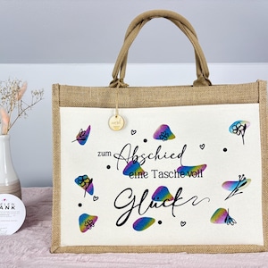 Pu&ograve; includere: Una borsa tote in iuta beige con un pannello frontale bianco. Il pannello frontale presenta motivi floreali colorati e il testo tedesco "zum Abschied eine Tasche voll Gl&uuml;ck". La borsa ha un manico beige abbinato.