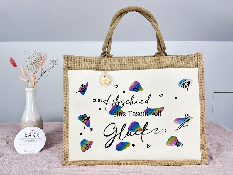 Pu&ograve; includere: Una borsa tote in iuta beige con un pannello frontale bianco. Il pannello frontale presenta motivi floreali colorati e il testo tedesco "zum Abschied eine Tasche voll Gl&uuml;ck". La borsa ha un manico beige abbinato.