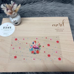 Könnte beinhalten: Dekorative Holzplatte mit einer Flamingo-Illustration, umgeben von Blumen. Auf der Platte steht der Text "amd AMBERGER DESIGN". Eine kleine Vase mit Trockenblumen und ein Aufkleber mit dem Text "VIELEN DANK" sind ebenfalls vorhanden.