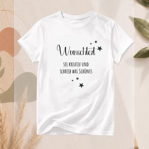 Könnte beinhalten: Ein weißes T-Shirt mit kurzen Ärmeln und schwarzem Text, der "Wunschtext" in einer Schreibschrift und "SEI KREATIV UND SCHREIB WAS SCHÖNES" in einer serifenlosen Schrift zeigt, verziert mit mehreren kleinen schwarzen Sternen.