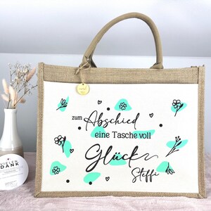 Pu&ograve; includere: Una borsa tote rettangolare con un pannello frontale bianco con scritte nere e accenti turchese. La borsa ha un telaio e manici in stile iuta, con un piccolo tag dorato attaccato. Il testo sulla borsa recita "zum Abschied eine Tasche voll Gl&uuml;ck Steffi."