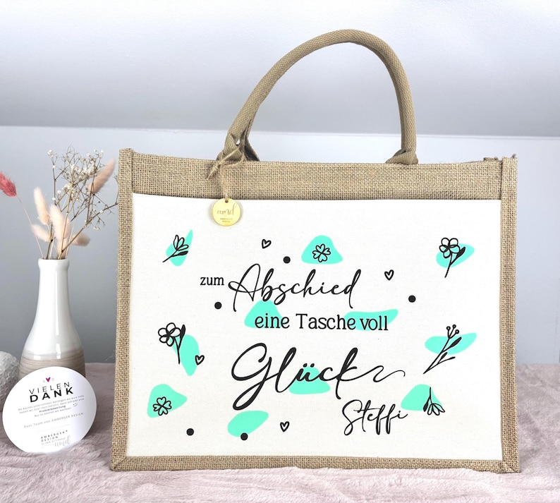 Pu&ograve; includere: Una borsa tote rettangolare con un pannello frontale bianco con scritte nere e accenti turchese. La borsa ha un telaio e manici in stile iuta, con un piccolo tag dorato attaccato. Il testo sulla borsa recita "zum Abschied eine Tasche voll Gl&uuml;ck Steffi."
